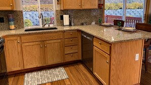 A kitchen with wooden cabinets, granite countertops, a dishwasher, and a window with a view of a stone wall and greenery. - Blackhawk 5BD Home: Hot Tub, Arcade, near Whitworth & Green Bluff (Spokane)
