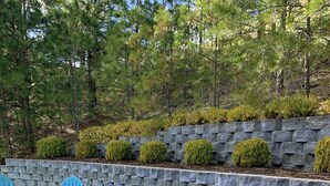 A hot tub surrounded by a stone wall and green bushes, with blue Adirondack chairs nearby. - Blackhawk 5BD Home: Hot Tub, Arcade, near Whitworth & Green Bluff (Spokane)