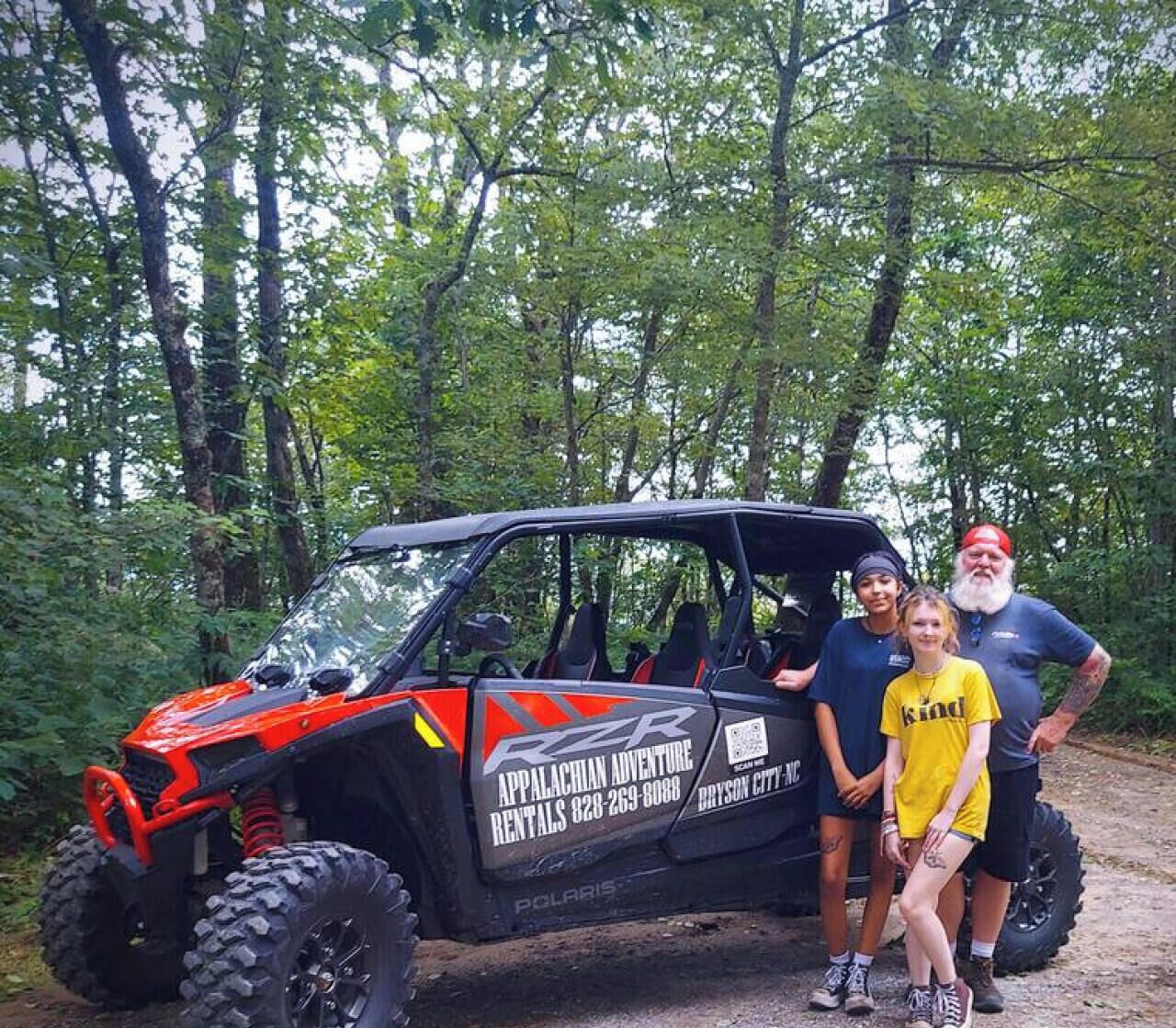Un véhicule Polaris RZR, orné d’une publicité pour une location, est garé sur un chemin de terre dans une zone boisée.