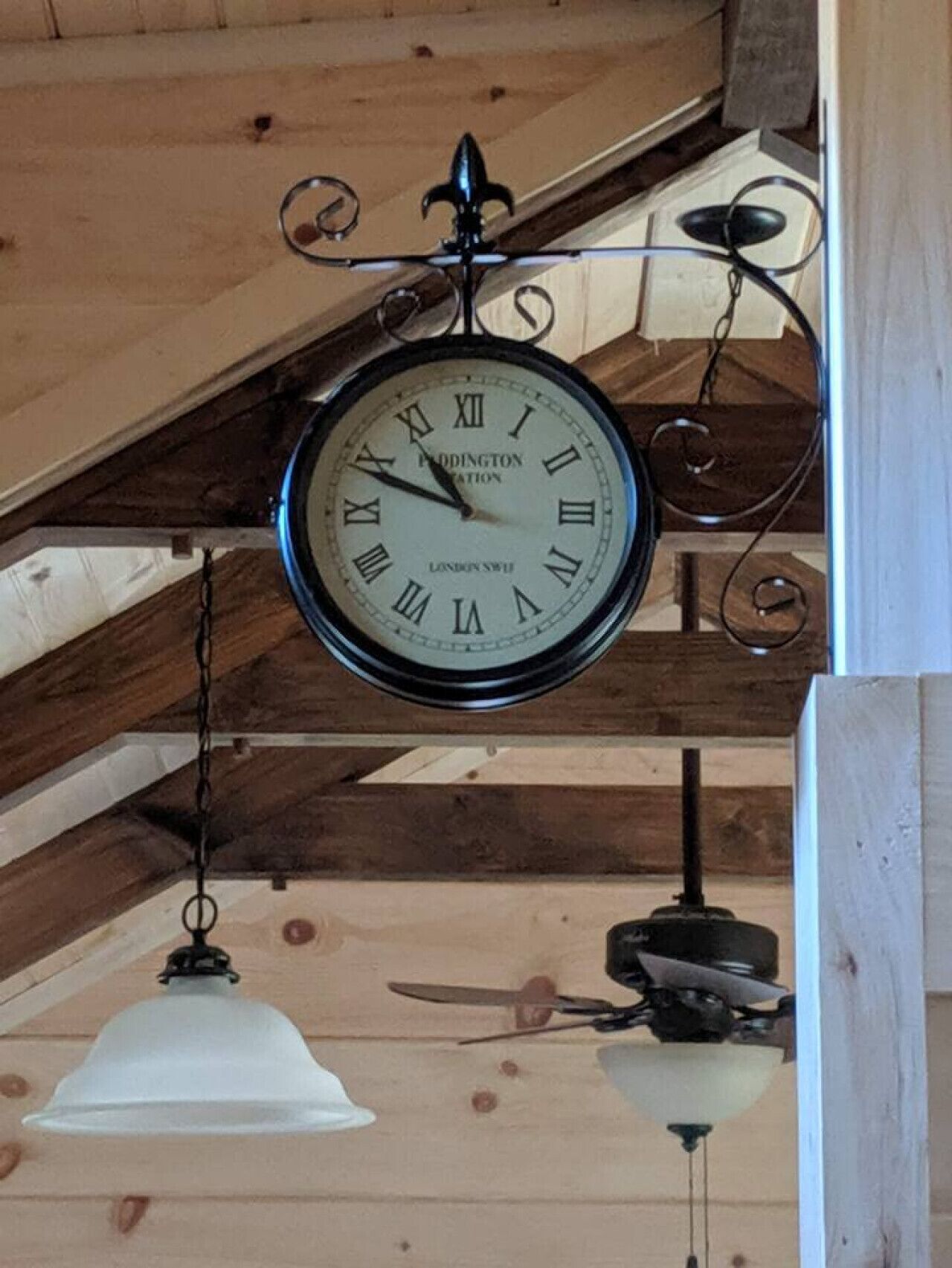 A wall-mounted clock with Roman numerals and the inscription 'LONDON SWIFT' in a room with wooden beams and a ceiling fan.