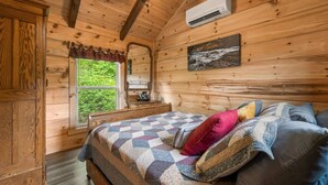 Quarto de cabana de madeira com uma cama, almofadas, um quadro, uma janela e um aparelho de ar condicionado.