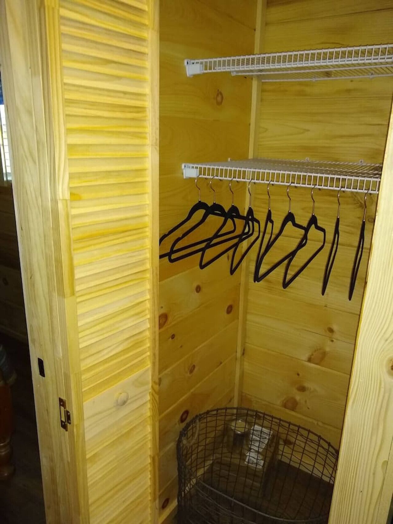 Quarto de madeira com uma estante de metal contendo vários cabides pretos, uma pequena gaiola de metal e uma prateleira branca.