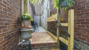 Un étroit sentier en briques mène à un portail en bois, avec un pot de fleurs et des meubles d’extérieur placés sur le côté gauche.