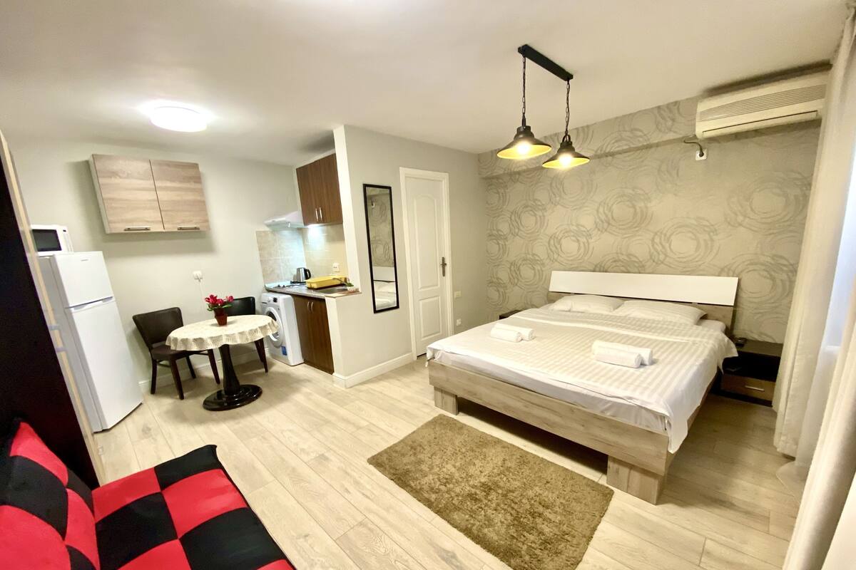 A bedroom with a bed, a small table with a chair, a washing machine, and a kitchenette.