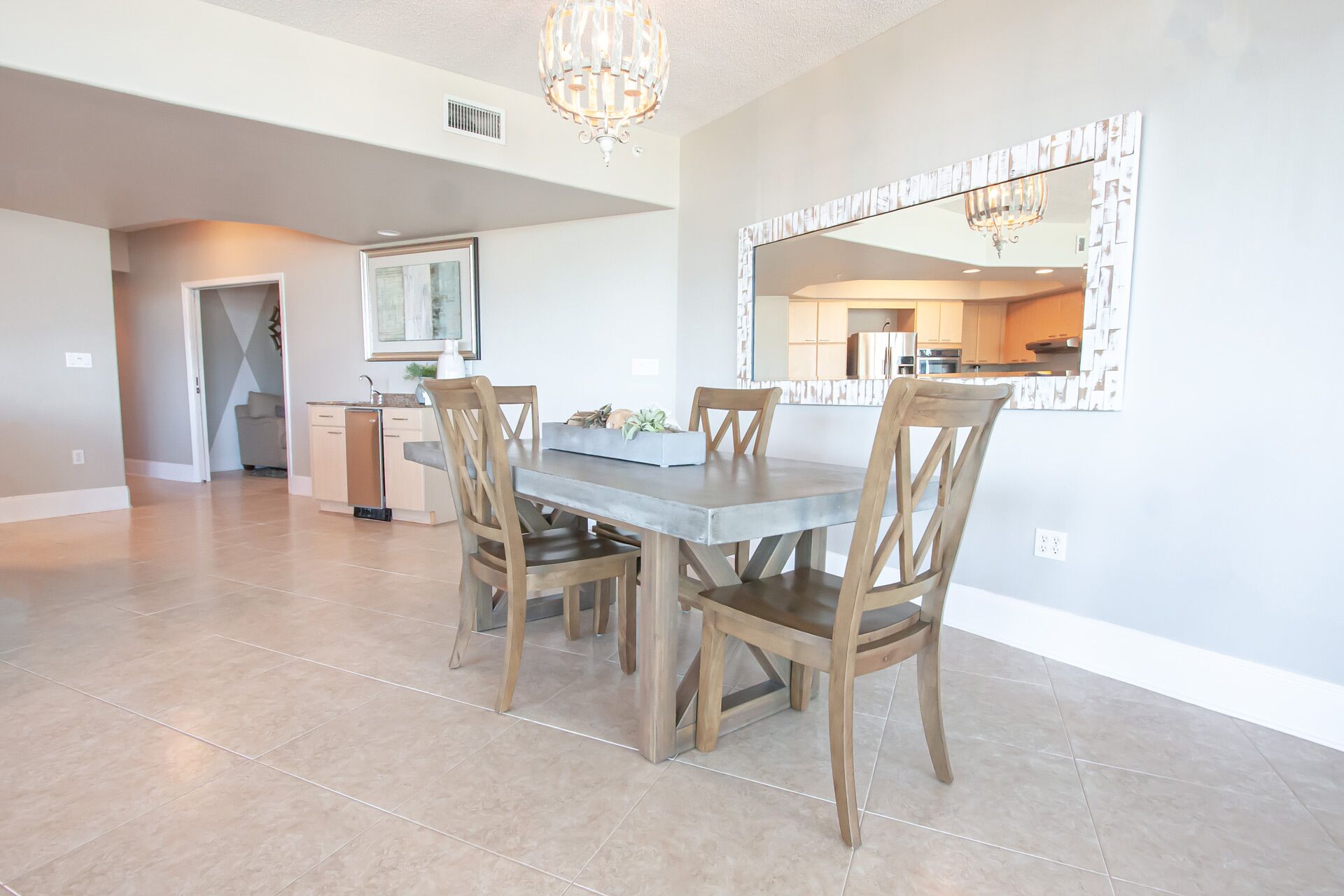 A dining area with a table and chairs, a kitchen in the background, and a large mirror on the wall.
