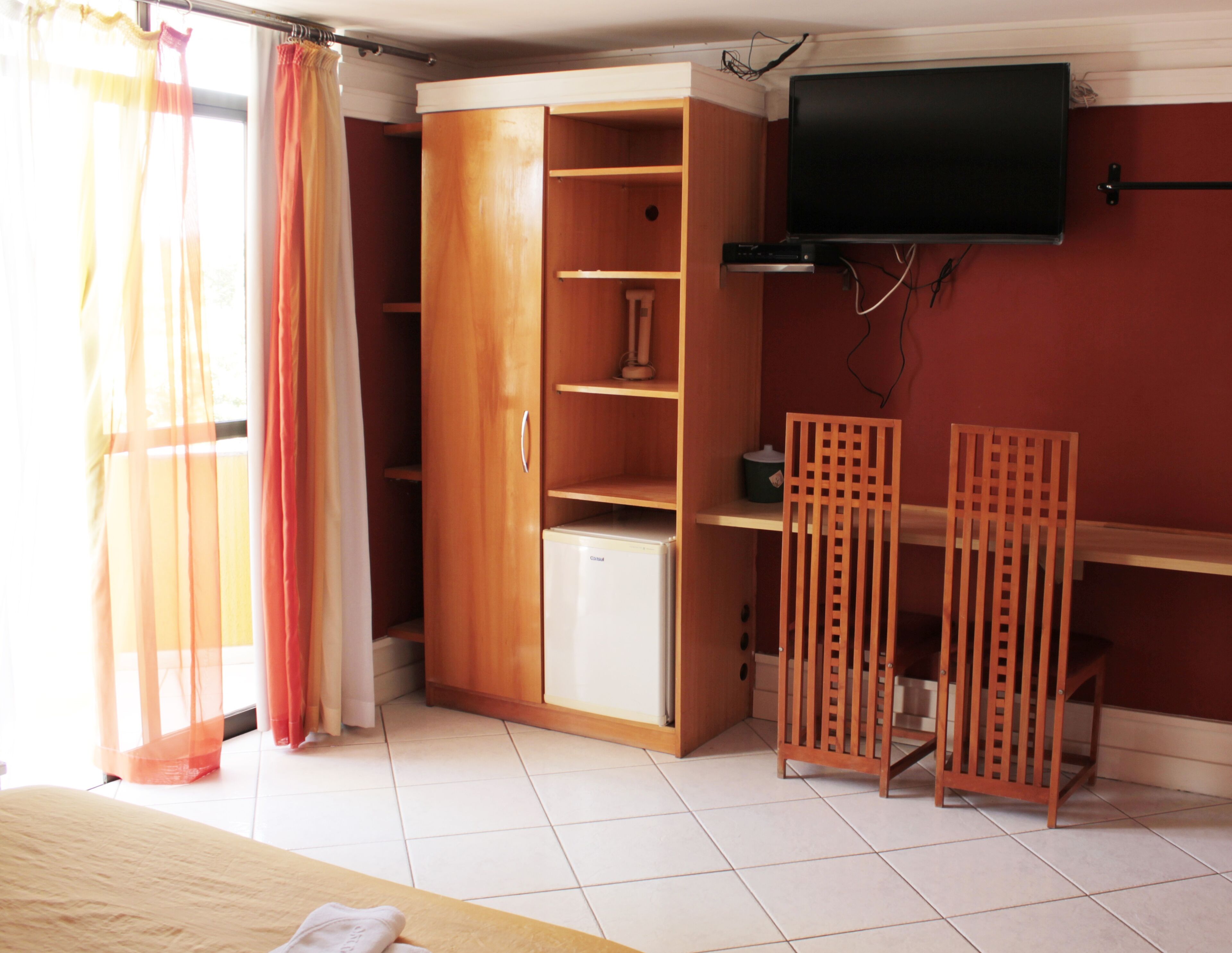 Quarto com guarda-roupa, um frigorífico pequeno, televisão na parede e uma cadeira de madeira.