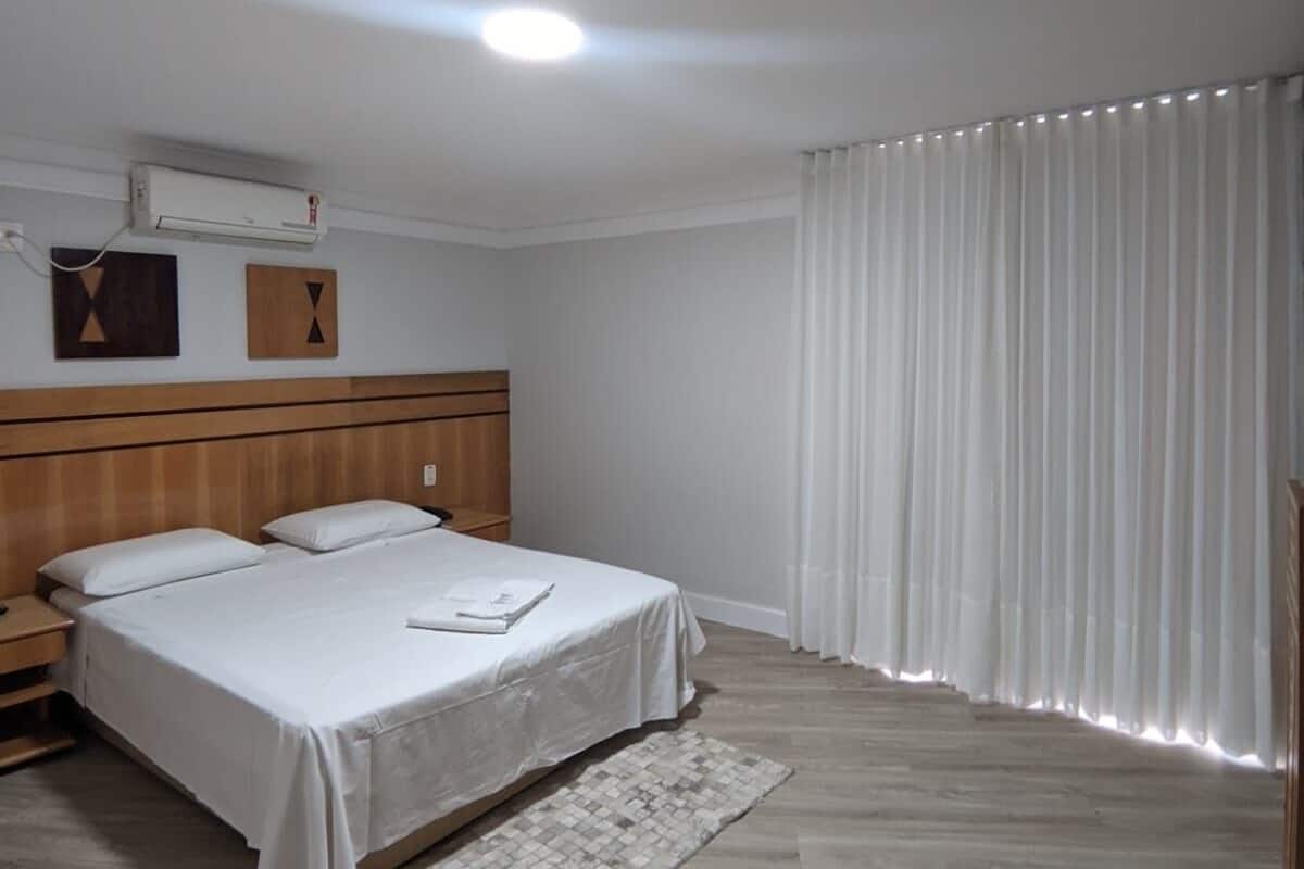 Quarto com cabeceira de madeira, cama com roupa de cama branca, mesa de cabeceira, tapete pequeno e janela com cortinas brancas.