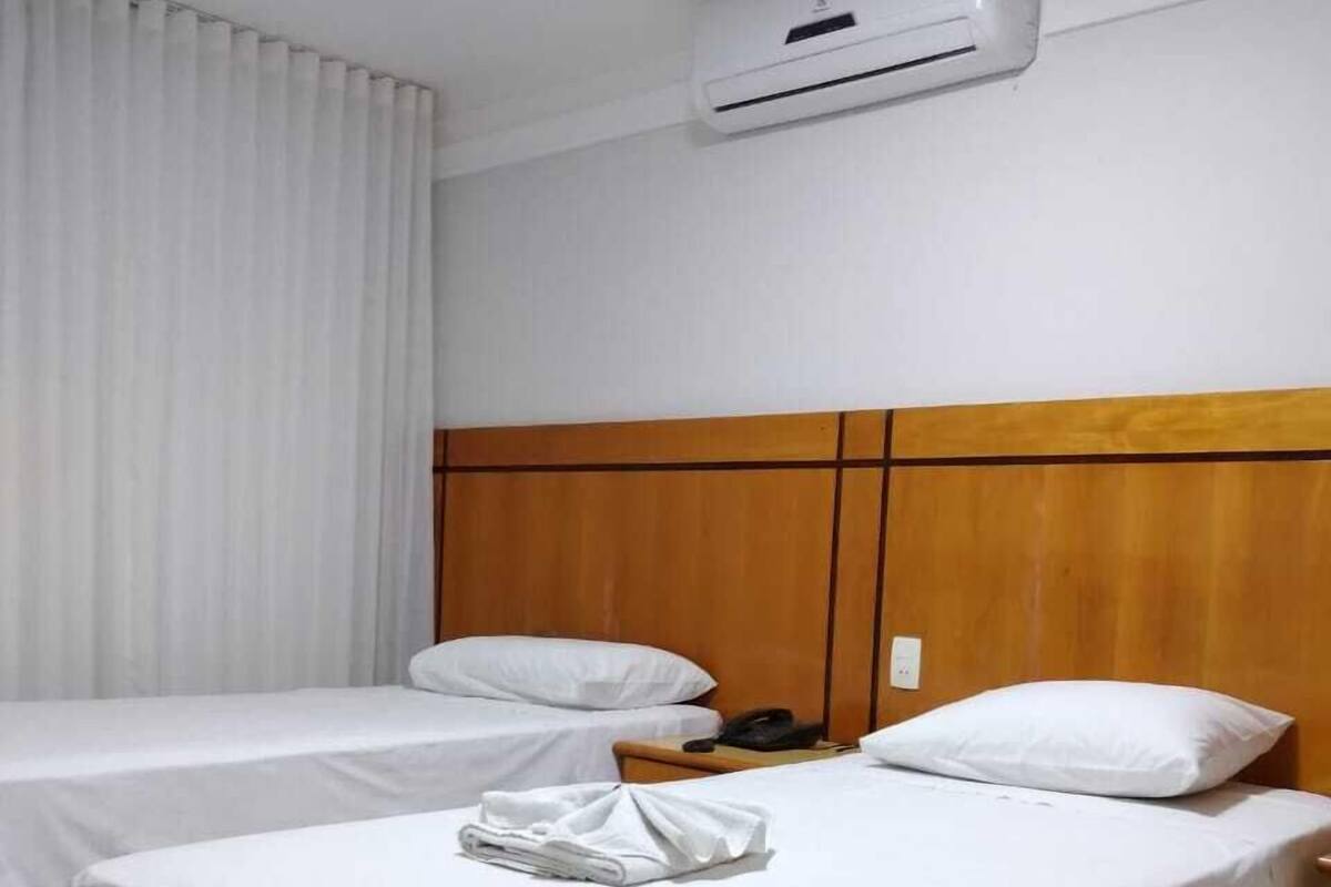 Quarto de hotel com duas camas de solteiro, roupa de cama branca, cabeceiras de madeira, ar condicionado branco e uma pequena mesa de cabeceira com telefone.