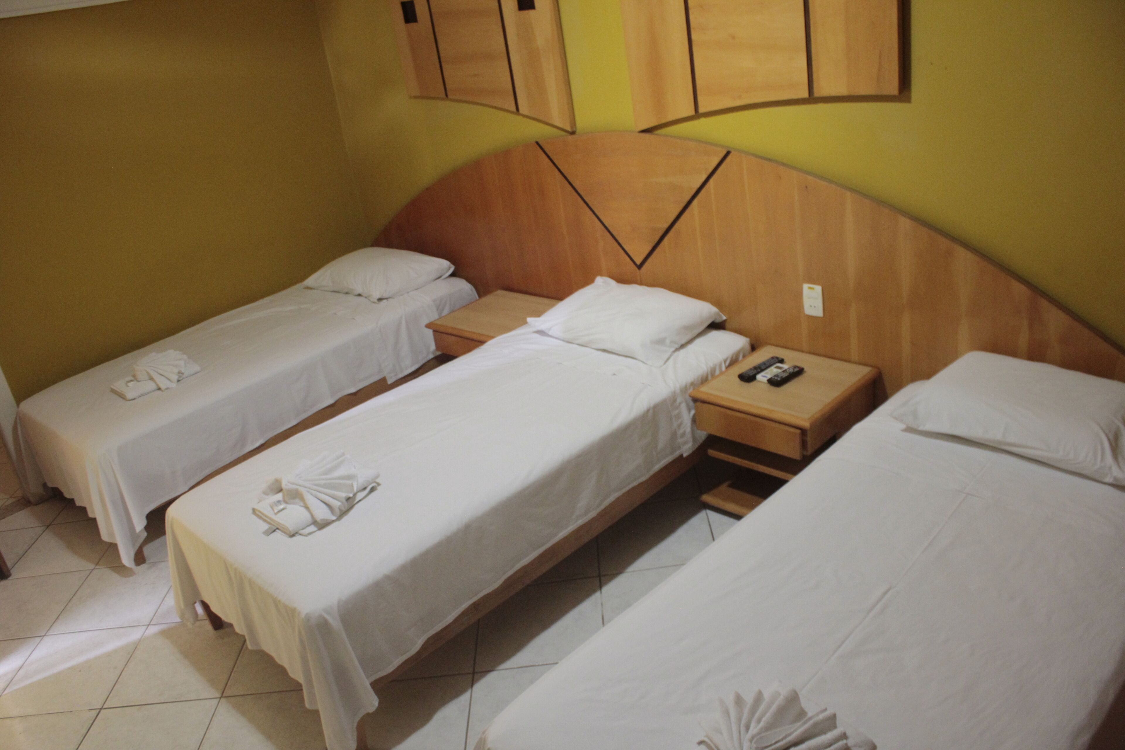 Quarto de hotel com três camas de solteiro, cada uma com roupa de cama e toalhas brancas. Possui cabeceira de madeira com design único e mesa de cabeceira de madeira com controle remoto.