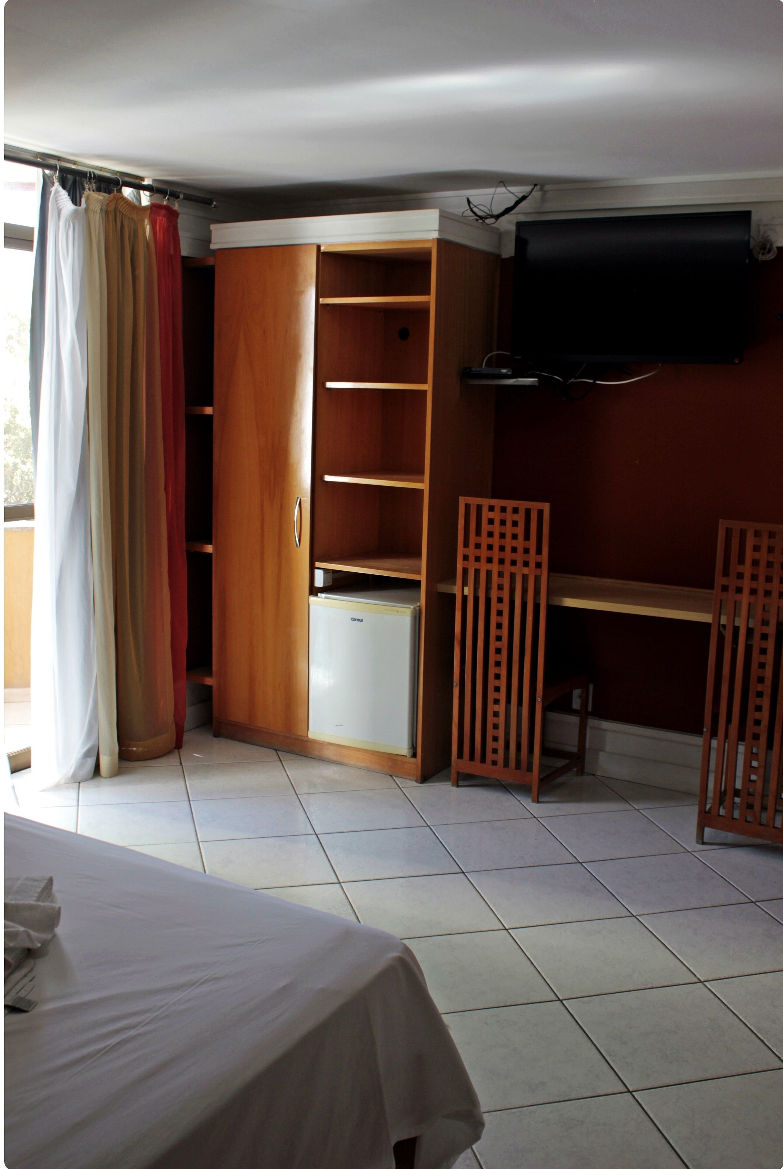 Una stanza con un letto, un'armadietto in legno, un piccolo frigorifero bianco e una TV fissata al muro.