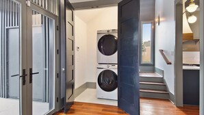 Une buanderie équipée de deux machines à laver et deux sèche-linge superposés, d’un escalier en bois et d’une porte donnant sur une autre pièce.