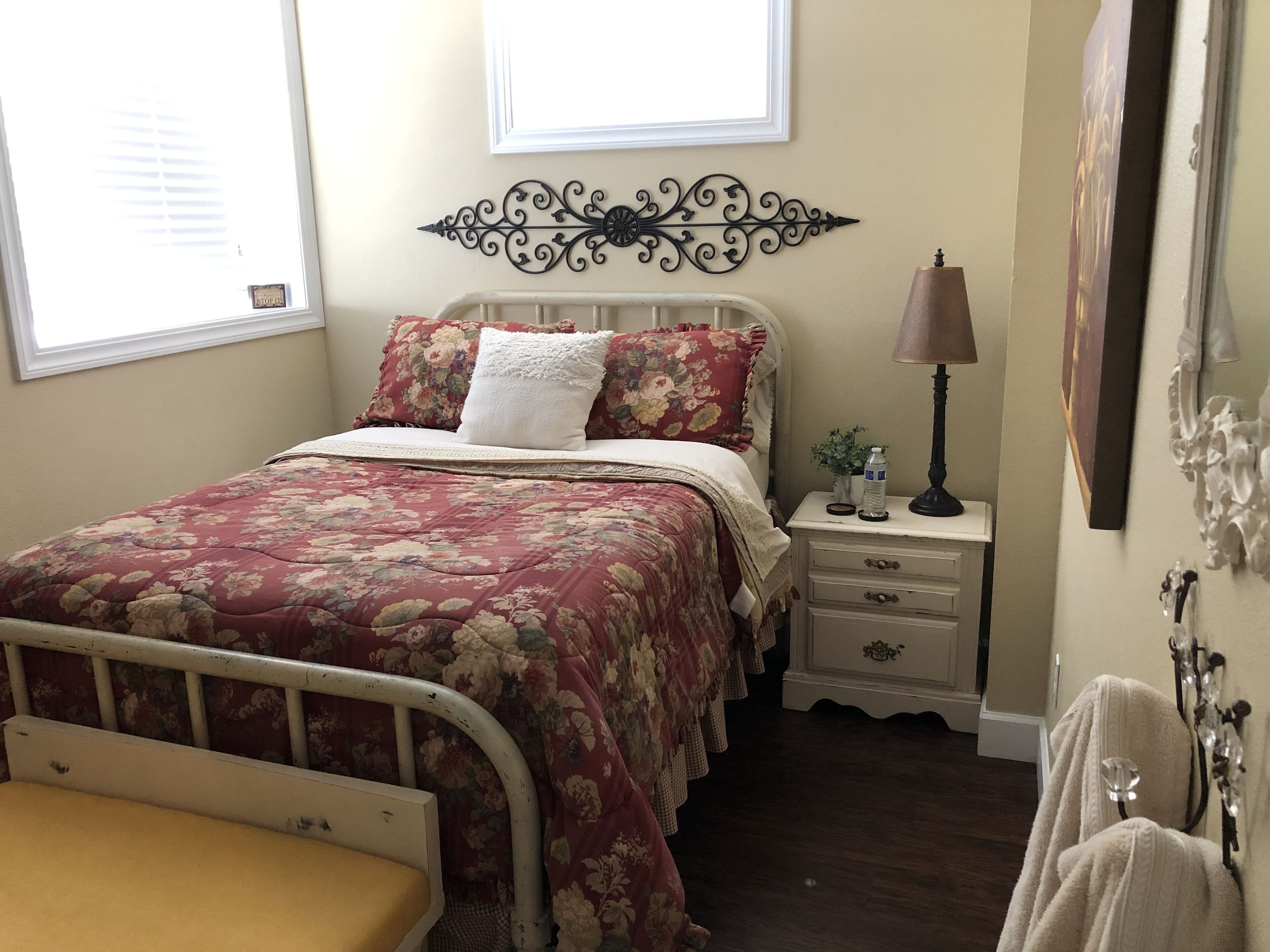 Une chambre à coucher avec une couvre-lit à motifs floraux, une tête de lit blanche, une table de chevet avec une lampe et un élément décoratif mural.