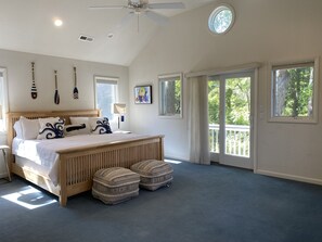 A bedroom with a large bed, wooden headboard, and two ottomans. There are multiple windows providing natural light and views of greenery outside. - Sound front Sunsets from Dock, Pool, Porch & all rooms. Private Beach access!  (Southern Shores)