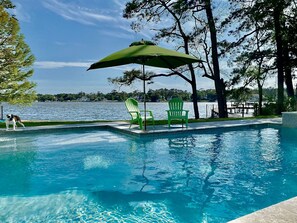 A swimming pool with clear blue water, surrounded by green chairs and an umbrella, with a view of a lake and trees in the background. - Sound front Sunsets from Dock, Pool, Porch & all rooms. Private Beach access!  (Southern Shores)