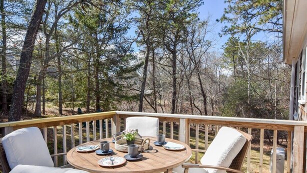 A wooden deck with a round table set for dining, surrounded by white cushioned chairs.