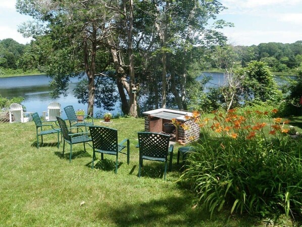 Un espace extérieur verdoyant avec une table et des chaises, une petite construction en briques et un lac en arrière-plan.