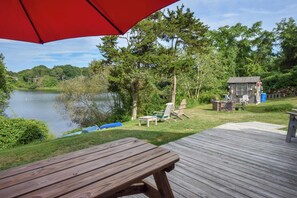 A wooden deck with a picnic table and chairs, overlooking a lake with trees and a small cabin. - Dream Waterfront A-Frame in Orleans! (Orleans)