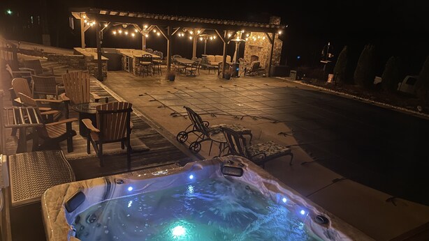 Un bain à remous entouré de pierres, un espace extérieur aménagé avec des chaises et des tables, et une cheminée en pierre située sous une terrasse couverte.