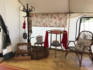 Um quarto com tenda, um cesto de vime, uma caixa de vime, uma cadeira de vime, uma mesa com um bule e uma chaleira, e um cabideiro com uma peça de roupa vermelha.