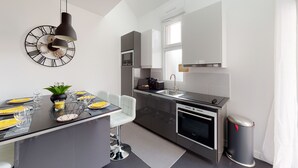 A modern kitchen with a dining area, featuring a black pendant light, a clock with Roman numerals, a stainless steel range hood, and a trash can. - Paris House (Villejust)