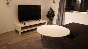 Un salon moderne avec un téléviseur à écran plat posé sur un meuble blanc, une table ronde blanche, un canapé marron et une plante en pot.