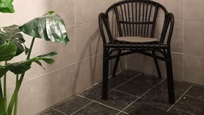 Un coin de pièce aux murs recouverts de carreaux beiges et au sol carrelé foncé. Une chaise noire est placée contre le mur, et une plante verte est visible sur la gauche.