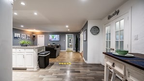 A modern kitchen with white cabinetry, a wooden floor, and a dining area with a dark wood table. - Mega House: 7 bed 3 bath! King & Queen beds! (Irving)