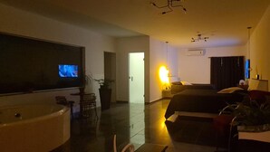 Habitación de hotel moderna con un televisor de pantalla plana grande, un sofá, una mesa de comedor y una planta grande en maceta.
