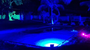 Escena nocturna con un área de piscina iluminada por luces azules y rosas, rodeada de palmeras y un muro de piedra.