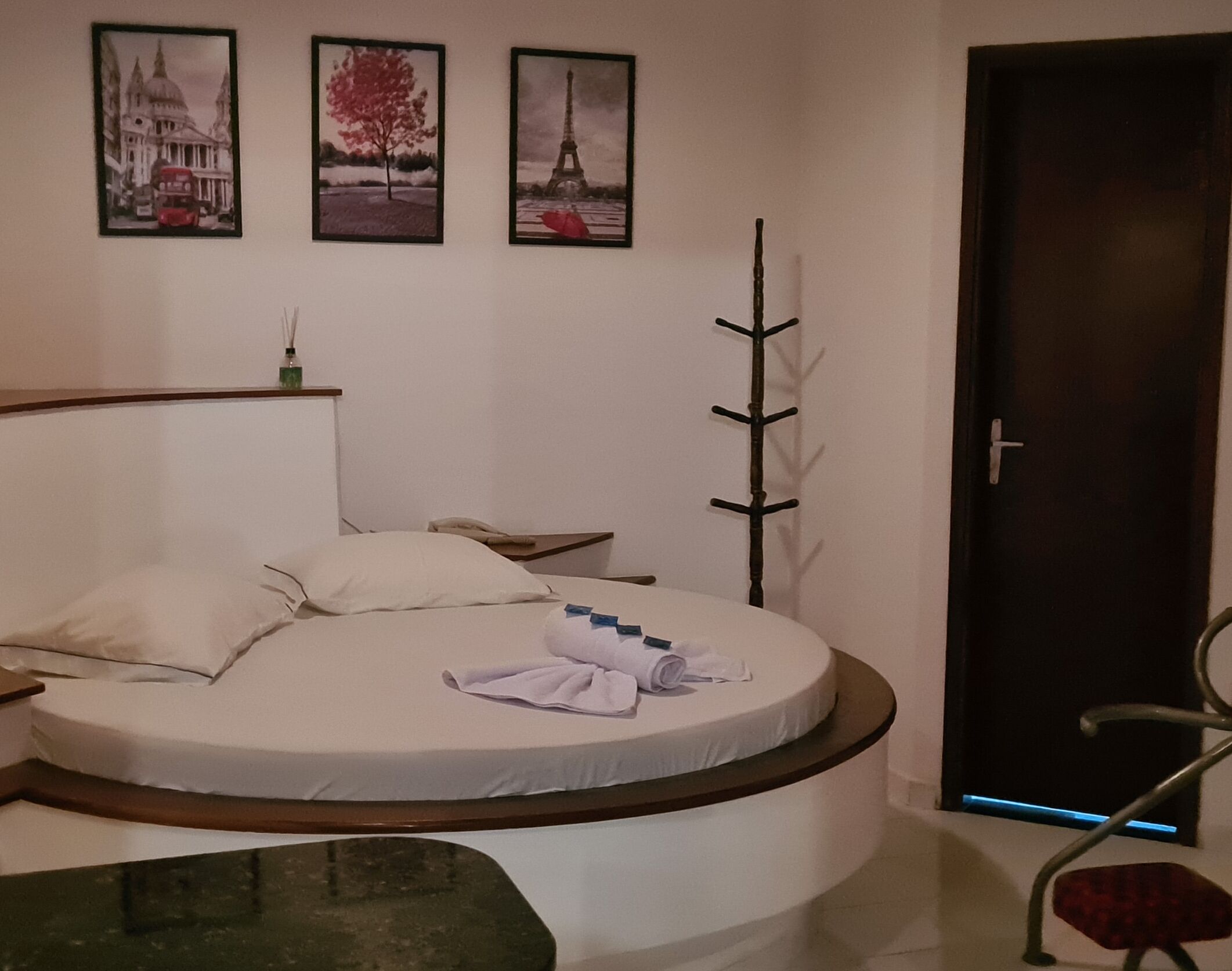 A circular bed with a wooden headboard, a bedside table, and a chair with a red cushion. There are three framed pictures on the wall above the bed.