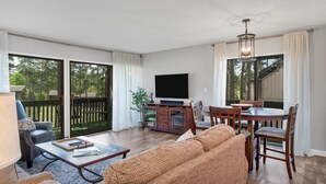A spacious living room with a large TV, a dining area, and a sliding glass door leading to a deck. - Pinseeker, a greenside condo on No. 5 - Walk to Pinehurst Clubhouse (Pinehurst)