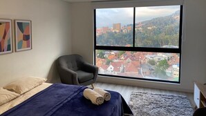 Una camera da letto con un letto, una sedia e un'ampia finestra vista città.