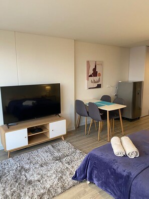 Ein modernes Hotelzimmer mit Flachbildfernseher, einem Esstisch mit Stühlen, einem Kühlschrank, einem Bett mit violetter Decke und einem grauen Teppich.