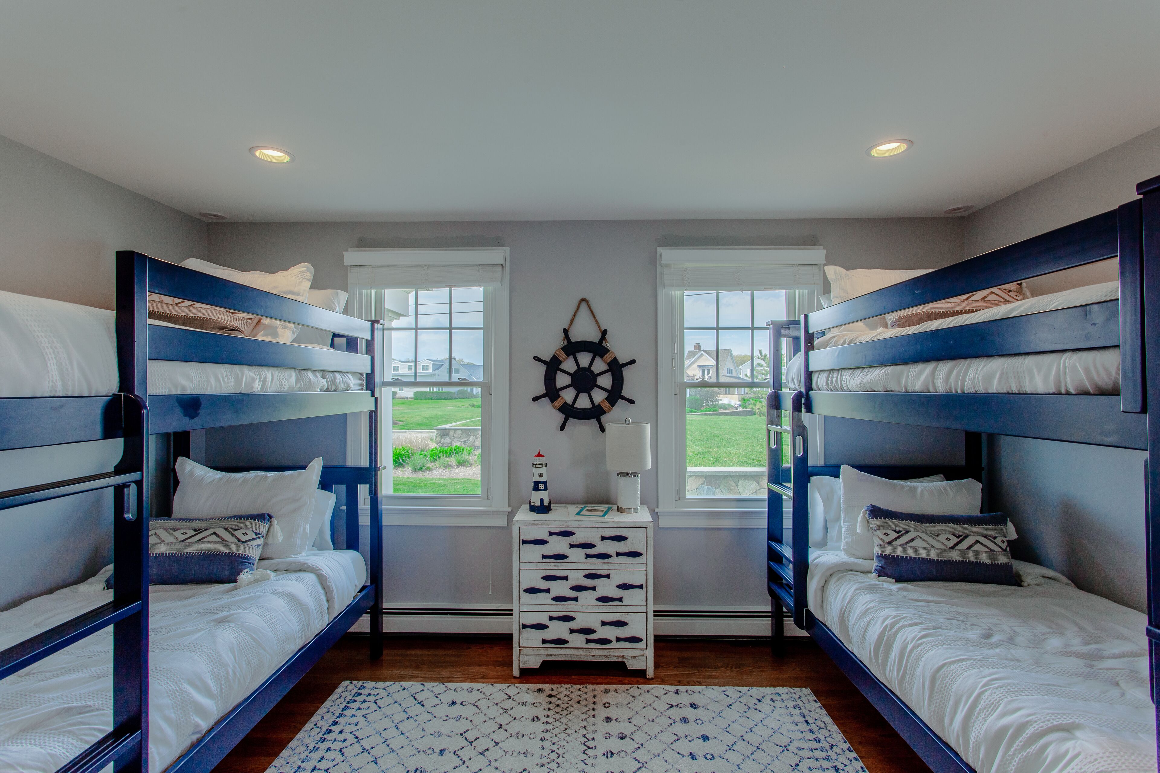 A room with two bunk beds, a small white nightstand with a ship's wheel decoration, and a view of a green landscape through two windows.