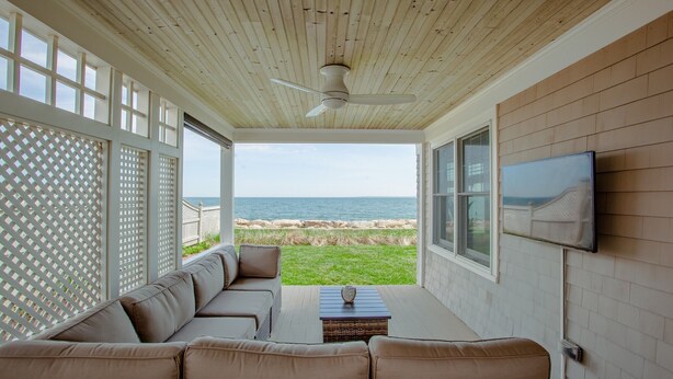 A covered outdoor seating area with a view of the ocean, a ceiling fan, and a flat-screen TV on the wall.