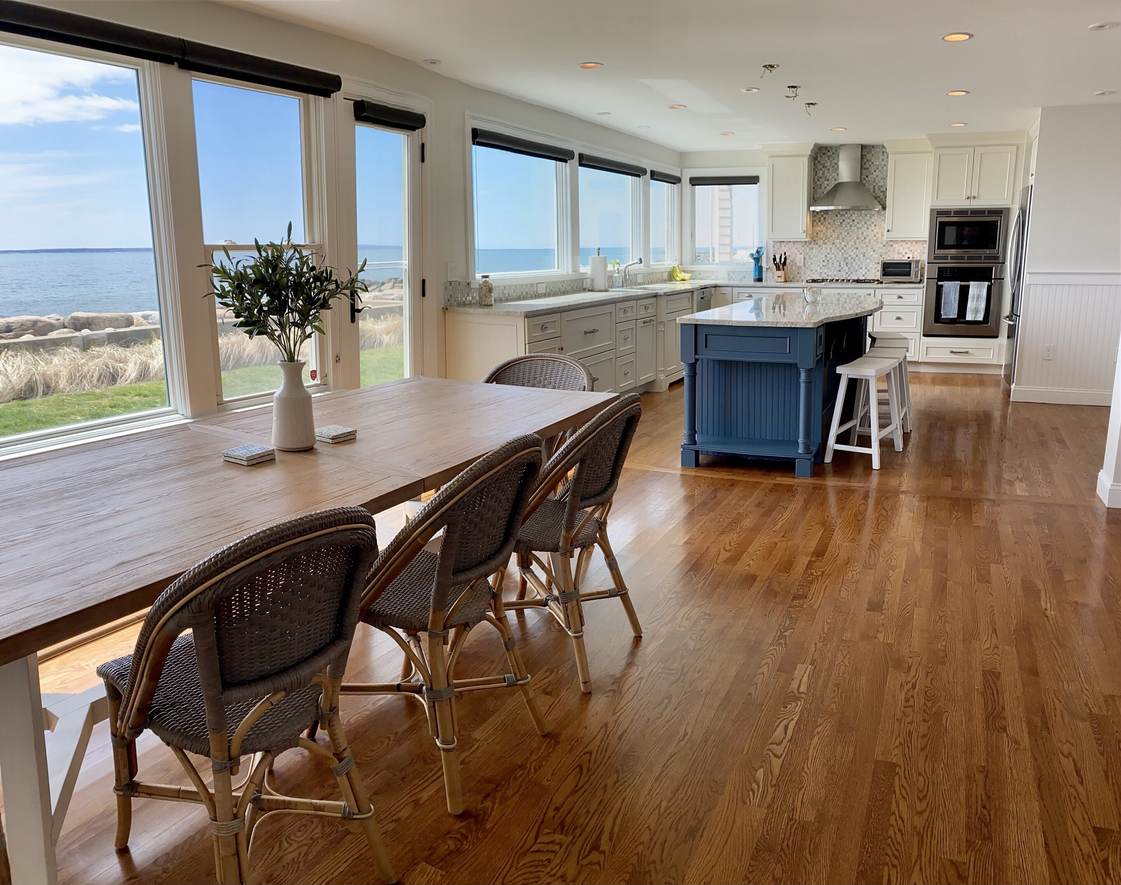 A modern kitchen with large windows, a wooden dining table, wicker chairs, and a view of the ocean.