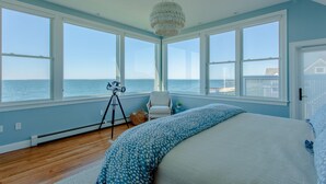 A bedroom with a large bed, a chair, a tripod, and a view of the ocean through large windows.