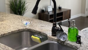 Eine Küche mit einer Spüle aus Edelstahl, einer Arbeitsplatte aus Granit, einem schwarzen Wasserhahn, einer kleinen Glasschale, einer grünen Flasche und einer Pfeffermühle.