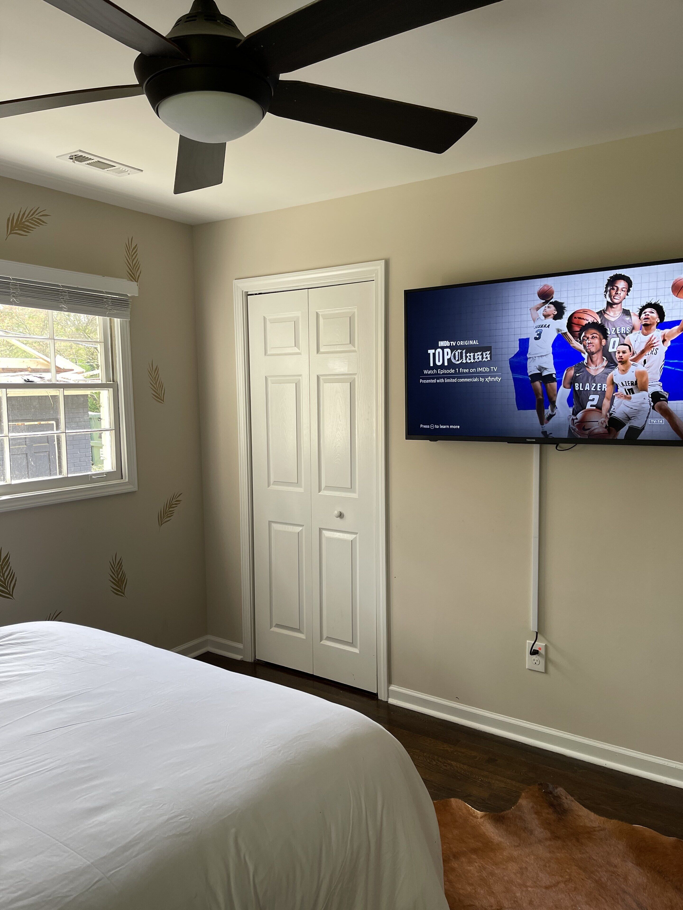 A bedroom with a bed, a ceiling fan, a television displaying a sports show, and a window with blinds.