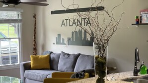 Ein modernes Wohnzimmer mit grauem Sofa, gelben Kissen, einem Glastisch und einer Küchenzeile mit Granit Arbeitsplatte und Spüle.
