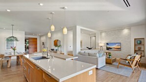A modern kitchen with a large island, wooden cabinets, pendant lighting, and a view of the living area with a sofa and a TV. - Maui Resort Rentals: Luana Garden Villas 15B – Modern 3BR Villa at Kaanapali’s Newest Luxury Residences! (Lahaina)
