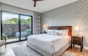 Quarto com uma cama grande, um ventilador de teto, uma mesinha de cabeceira com abajur e vista para o verde através de uma porta de vidro deslizante.