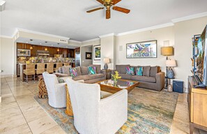 A modern living room with a sofa, coffee table, and chairs, a kitchen with wooden cabinets, and a ceiling fan.