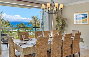 A dining area with wicker chairs and a table set for six, overlooking a beach with palm trees and the ocean.
