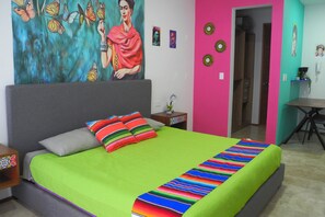 A bedroom with a large bed, colorful pillows, a vibrant painting on the wall, and a small table with a plant.