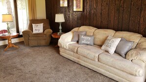 Un salon chaleureux avec un mur en bois brun, un canapé beige orné de coussins à motifs, un fauteuil brun, une petite table d’appoint en bois et une lampe.
