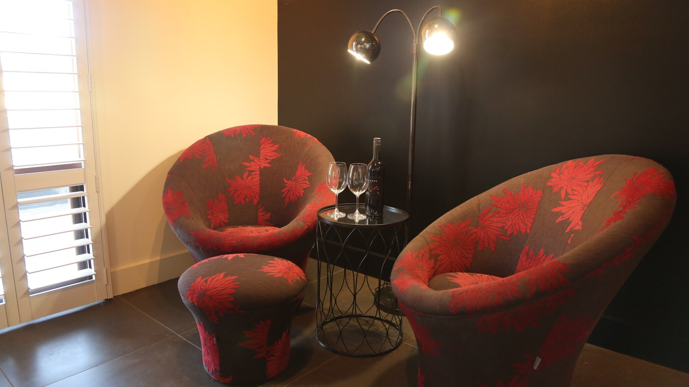 Una habitación con dos sillones tapizados con motivos de hojas rojas, una mesita redonda con una botella y copas, y una lámpara de pie.