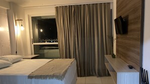 A hotel room with a bed, a TV mounted on the wall, a small table, and a window with curtains. - Studio Business West Flat (Mossoró)
