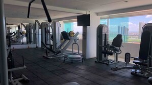 A gym with various exercise machines, a bench, and a large window offering a city view. - Studio Business West Flat (Mossoró)