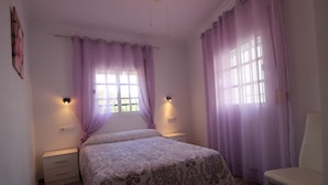 A bedroom with a bed, two bedside tables, a chair, and a window with purple curtains. - Apartment in Conil de la Frontera, Cádiz (Conil de la Frontera)